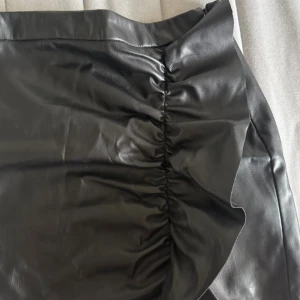 Svart skinnkjol  - Snygg svart kjol i skinnimitation med rynkade detaljer längs sidan och en vågig volang. Kjolen har en tight passform 