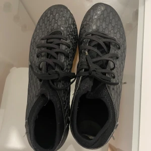 Svarta fotbollsskor med mönster - Säljer ett par svarta fotbollsskor med coolt prickigt mönster och snörning. Skorna har rund tå och platt sula med dobbar för bra grepp på planen. Ovandelen är i syntetmaterial och insidan är mjuk för komfort. Perfekt för dig som vill sticka ut på fotbollsplanen.