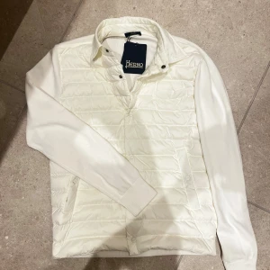 Vit quiltad pufferjacka från Herno - Snygg vit pufferjacka från Herno med quiltad framsida och släta stickade ärmar och rygg. Jackan har krage, knappar framtill och två sidofickor. Perfekt för dig som gillar stilren design och vill ha en jacka som sticker ut med sin mix av material.