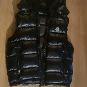 Moncler väst - Snygg svart dunväst från Moncler med glansig finish och klassisk logga på bröstet. Västen har quiltad design, dragkedja framtill och två fickor. Perfekt för lager-på-lager och streetstyle. Fodrad med dun för extra värme.