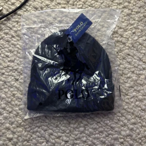 Svart mössa från Polo Ralph Lauren - Svart ribbstickad mössa från Polo Ralph Lauren med klassisk broderad logga i vitt framtill. Mjuk och varm, perfekt för kyliga dagar. Stilren design med uppvikt kant och diskret tagg. Passar dig som gillar enkel och snygg streetstil.