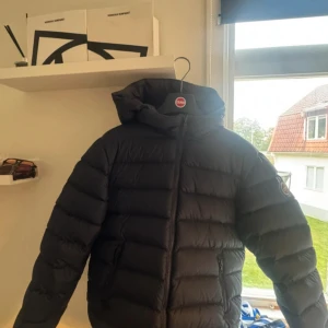 Mörkblå Colmar pufferjacka med huva - Säljer en mörkblå pufferjacka från Colmar med stor huva och tydlig logga på ärmen. Jackan har dragkedja framtill och är quiltad för extra värme. Perfekt för dig som vill ha en stilren och varm jacka under kalla dagar. Har en liten defekt på under arm men som ingen har lagt märke till och man inte märker.