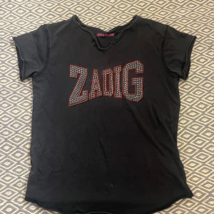 Svart t-shirt från Zadig & Voltaire - Snygg svart t-shirt från Zadig & Voltaire med stort ZADIG-tryck i vita och röda prickar på framsidan. T-shirten är kortärmad och tillverkad i 100% bomull. Finns ingen storlek men är cirka xs.