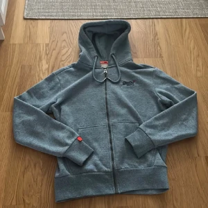 Blå Superdry hoodie med dragkedja - Blå hoodie från Superdry Orange Label med broderad logga på bröstet. Tröjan har huva med snörning, hel dragkedja och två fickor framtill. Mjuk och skön bomullsblandning, perfekt för chill dagar eller när du vill ha en clean streetstyle.