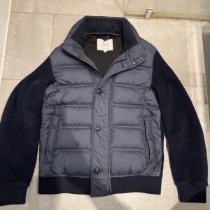 Svart pufferjacka från Woolrich - Snygg mörkblå pufferjacka från Woolrich i storlek S. Jackan har quiltad front med både dragkedja och knappar, samt stickade ärmar och muddar. Hög krage och två sidofickor ger en clean och modern look. Perfekt för dig som gillar stilren och funktionell design. Jackan håller även värmen bra då den är fodrad  med varm ull som gör att man håller sig varm under en kall vinter dag. Jackan är i mycket bra skick och använd endast 2 månader. Jackan köptes för runt 4500kr-5000kr. Hör av er vid frågor😊