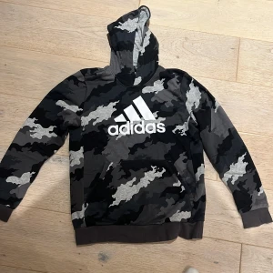 Svart camo Adidas hoodie med logga - Säljer en svart och grå camo hoodie från Adidas med stor vit logga på bröstet. Tröjan har huva, känguruficka och är långärmad. Perfekt för dig som gillar streetstyle och sportig vibe. Barn storlek.