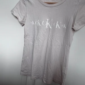 Beige t-shirt från Calvin Klein Jeans - Snygg beige t-shirt från Calvin Klein Jeans med vit CK-logga tryckt flera gånger över bröstet. Klassisk rund halsringning och korta ärmar. Tillverkad i mjuk bomull som är skön mot huden. Oanvänd men prislappen är borta. SKRIV INNAN KÖP!