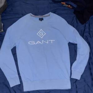 Ljusblå sweatshirt från GANT - Snygg ljusblå sweatshirt från GANT i storlek M. Tröjan har ett stort vitt GANT-tryck och logga på bröstet, rund hals och ribbade muddar vid ärmar och nederkant. Tillverkad i mjuk bomull, perfekt för chill dagar och enkel att matcha med jeans.
