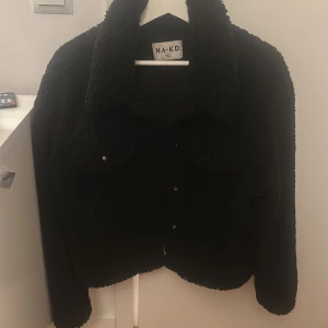 Svart teddyjacka från NA-KD - Mysig svart teddyjacka från NA-KD med stora fickor framtill och krage. Jackan har knappar och är perfekt för att hålla värmen under kyliga dagar. 