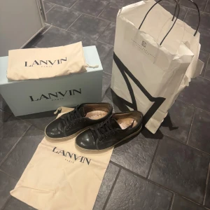 Svarta sneakers från Lanvin i skinn och mocka - Snygga svarta sneakers från Lanvin med ovandel i både skinn och mocka. Klassisk rund tå, svarta snören och vit sula. Kommer med originalkartong och dustbags. Perfekta för dig som gillar stilrena och exklusiva sneakers med lyxig känsla. Det finns digitalt kvitto från nk. Jag har också bytt snören till helt nya, de mesta av smutsen går bort ganska enkelt och det går att fixa mockan med mocka cleaning kit. Skorna passar för 42 också