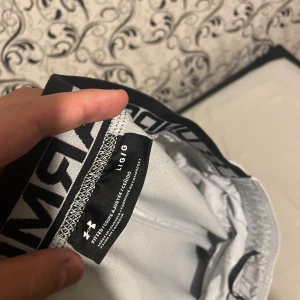 Ljusgrå träningsshorts från Under Armour - Snygga ljusgrå träningsshorts från Under Armour med elastisk midja och svart logotyp på vänster ben. Shortsen har en bred resår med tydlig branding och är tillverkade i ett lätt syntetmaterial som andas. Perfekta för gymmet eller löprundan.