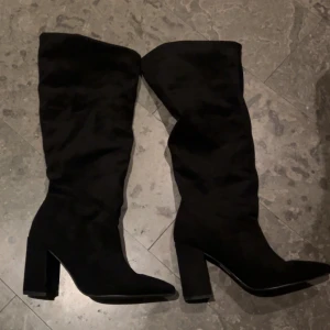 Svarta mocka stövlar med klack - Säljer ett par svarta höga stövlar med bred klack och spetsig tå. Stövlarna är tillverkade i mocka och har en stilren, elegant look som passar perfekt till jeans eller kjol. De är knähöga och har en plattformad sula för extra komfort.