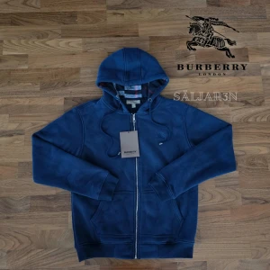 Burberry Hoodie - Snygg marinblå hoodie från Burberry med dragkedja, huva och klassiskt rutigt foder på insidan. Broderad Burberry-logga på bröstet och exklusiva metalldetaljer på snörningen. Tillverkad i mjuk bomull och polyesterblandning, perfekt för höst och vinter.