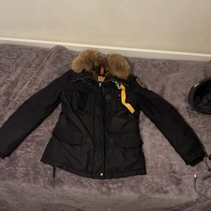 Svart Parajumpers parkajacka med päls - Säljer en svart Parajumpers Anchorage parkajacka med fluffig brun päls på kragen och insidan. Jackan har flera fickor, gul detalj vid dragkedjan och patch på ärmen. Perfekt för kalla dagar med sin varma och stilrena look.