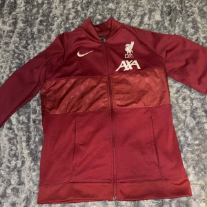 Liverpool FC träningsjacka Nike vinröd - Snygg vinröd träningsjacka från Nike med Liverpool FC-logga och AXA-tryck på bröstet. Jackan har hel dragkedja, två fickor och ett mönstrat band över bröstet och ryggen. Perfekt för dig som älskar fotboll och vill representera LFC.