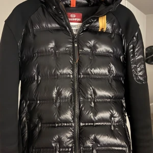 Svart pufferjacka från Parajumpers - Säljer en svart pufferjacka från Parajumpers med glansig yta och quiltad design. Jackan har huva, dragkedja framtill och en gul detalj vid halsen. Perfekt för kalla dagar och har en modern, sportig look. Storlek L sitter som M. 