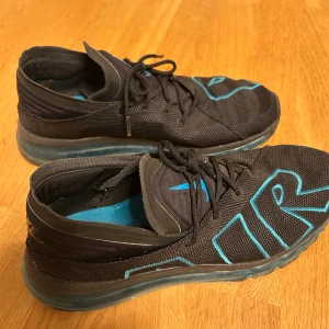 Nike Air Max sneakers svart/blå - Nike Air Max sneakers i svart med stora blå AIR-detaljer på ovansidan. Skorna har snörning, platt sula med synlig luftkudde och rund tå. Ovandelen är i syntetmaterial och mesh för bra ventilation. Perfekt för dig som gillar sportig och modern stil.