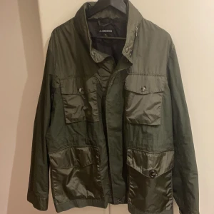 Grön parkajacka från J.Lindeberg - Snygg grön parkajacka från J.Lindeberg med coola detaljer och flera stora fickor framtill. Jackan har en hög krage, knappar och dragkedja samt ett metallmärke på ärmen. Materialet är polyester och bomull, perfekt för dig som gillar stilren design.