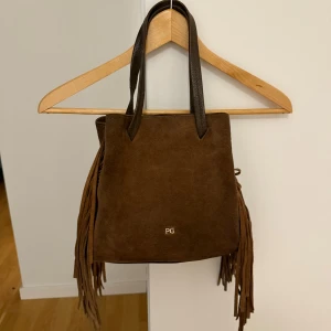 Brun mocka axelväska med fransar PG - Snygg brun axelväska i mocka från PG med långa fransar på sidorna och mörkbruna handtag i skinn. Väskan har en fyrkantig form och stilren design med diskret PG-logo i guld framtill. Perfekt för dig som vill ha en cool och trendig accessoar.