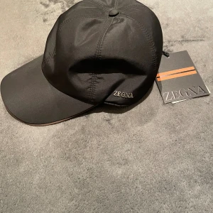 Svart keps från Zegna i storlek M - Snygg svart keps från Zegna med stilren design och metallogo framtill. Kepsen har en böjd skärm och insida med orange-randigt band. Materialet är slätt och känns lyxigt, perfekt för dig som gillar exklusiva accessoarer.