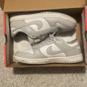 Nike Dunk Low vit och grå sneakers - Nike Dunk Low sneakers i en clean vit och ljusgrå färg. Klassisk siluett med rund tå, snörning och platt sula. Ovandel i skinn med textilfoder för extra komfort. Perfekt för dig som gillar stilrena och tidlösa sneakers.