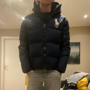 Mörkblå tjockjacka från Ralph Lauren - Snygg mörkblå dunjacka från Ralph Lauren med stor vit logga på bröstet. Jackan har huva, dragkedja framtill och två sidofickor. Puffad design som ger en cool siluett och håller dig varm under kalla dagar kontakta för fler frågor eller bilder !.