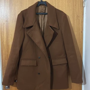 Brun skepparkavaj med bred krage - Snygg brun skepparkavaj med bred krage och dubbelknäppning framtill. Jackan har två stora fickor med lock och är helfodrad på insidan. Perfekt för dig som gillar klassisk stil med en modern twist.