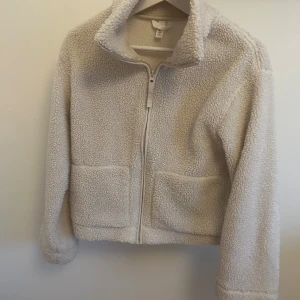 Beige teddyjacka  - Mysig teddyjacka i beige med hög krage och två stora fickor framtill. Jackan har hel dragkedja och är tillverkad i fluffigt fleece-material som ger en chill och varm vibe. Perfekt för lager-på-lager och passar dig som gillar en enkel, clean look. Kan tvättas innan vid önskan 