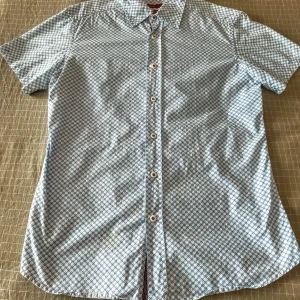 Mönstrad kortärmad skjorta Zara - Kortärmad skjorta från Zara i 100% bomull med ljusblått och vitt geometriskt mönster. Skjortan har klassisk krage, vita knappar och coola röda ränder på insidan av kragen. Perfekt för dig som gillar unika detaljer och snygg passform!