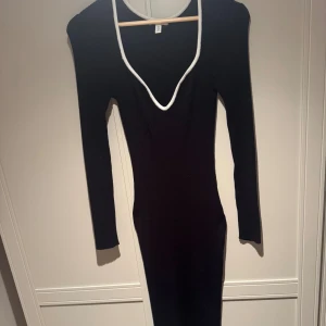 Svart ribbad långklänning med vit kant (Mama) - Supersnygg svart långklänning med ribbad struktur och vit kant runt den djupa v-ringningen. Klänningen har lång ärm och figurnära passform som framhäver siluetten. Perfekt för dig som gillar minimalistisk stil med en twist. (Det både en gravid/mamma klänning) använd vid 1 tillfälle. 