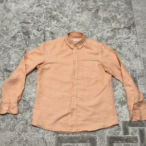 Persikofärgad linneskjorta från Zara - Persikofärgad skjorta från Zara i storlek S, tillverkad i mjukt linne. Klassisk modell med knappar framtill, bröstficka och button-down krage. Långärmad och med en clean look som är lätt att matcha med jeans eller chinos.