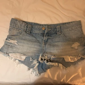 Blå jeansshorts med slitningar Gina Tricot - Snygga ljusblå jeansshorts från Gina Tricot med råa, fransiga kanter och slitna detaljer framtill. Klassisk femficksmodell med bälteshällor och knappstängning. Perfekta för sommaren och ger en cool, avslappnad vibe.
