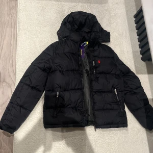 Svart pufferjacka från Polo Ralph Lauren - Svart pufferjacka från Polo Ralph Lauren med huva och två sidofickor med dragkedja. Jackan har det klassiska röda Polo-logot broderat på bröstet och quiltad design. Perfekt för kalla dagar och har en snygg, sportig look.
