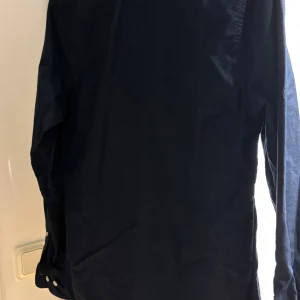 Mörkblå skjorta från East West - Snygg mörkblå skjorta från East West med klassisk button-down krage och vita knappar hela vägen. Skjortan har lång ärm och är tillverkad i mjuk bomull. Perfekt för dig som gillar stilrena och enkla plagg med en clean look. Storlek xs, men sitter som S 