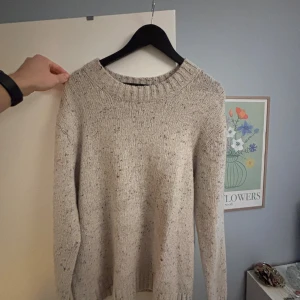 Beige stickad tröja från Sandö - Säljer en beige stickad tröja från Sandö i storlek L. Tröjan har ett grovt ribbstickat parti vid halsen och är dekorerad med små färgstänk i garnet för en cool, lite vintage look. Perfekt för dig som gillar enkel och stilren design med lite extra detaljer.