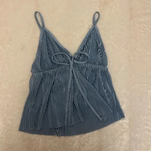 Blå randig linnetopp med knyt - Supersöt blå linnetopp med tunna axelband och v-ringad hals. Toppen har ett randigt mönster och knyt framtill för extra detalj. Materialet är mjukt och lätt, perfekt för varma dagar. Okänt märke och storlek men uppskattar storlek Xs