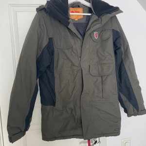 Grön parkajacka från Fjällräven XS - Parkajacka från Fjällräven i mörkgrön och svart färg, med huva och flera praktiska fickor framtill. Jackan har dragkedja och knappar, samt Fjällräven-logga på bröstet. Perfekt för kalla dagar och outdoor-häng.
