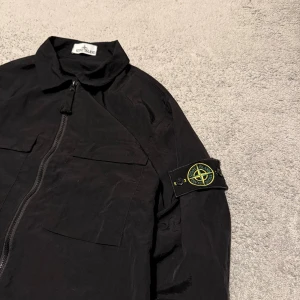 Svart overshirt från Stone Island - Exklusiv Stone Island overshirt i svart – en riktig ikon inom streetwear och premium casual mode. Tillverkad i högkvalitativt material med den klassiska Stone Island-kompassen på ärmen. Perfekt som lätt jacka eller layering-plagg året runt.  ✅ Märke: Stone Island ✅ Storlek: L ✅ Färg: Svart ✅ Skick: Mycket bra – sparsamt använd, inga defekter eller missfärgningar ✅ Detaljer: Avtagbart märke på ärmen, dragkedja framtill, två stora bröstfickor ✅ Stil: Modern, trendig & exklusiv 