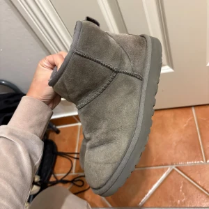 Grå UGG boots i mocka, ÄKTA! - Säljer ett par klassiska UGG boots i grå mocka med rund tå och platt sula. Insidan är fodrad med mjuk päls som håller fötterna varma. Bootsens design är enkel och tidlös, med UGG-logga baktill och grov yttersula för extra grepp.  De är tyvärr ej lika fkuffiga vid häl och hälrygg men inga större defekter än så. Köpta i raglady på väla för 2000kr.Original låda tillkommer!!
