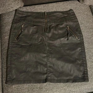 Svart kortkjol med dragkedjor - Snygg svart kortkjol med coola detaljer som dragkedjor framtill och fickor både fram och bak. Kjolen har en slim passform och är gjord i ett syntetmaterial med lätt glansig finish. Perfekt för dig som gillar en edgy stil och vill ha något som sticker ut.