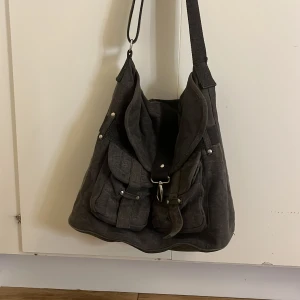 Grå axelväska från b.p.c i bomull - Snygg grå axelväska från b.p.c i 100% bomull. Väskan har två framfickor med lock och spännen, justerbar axelrem och ett stort huvudfack med dragkedja på insidan. Perfekt för dig som vill ha plats för allt du behöver under dagen.