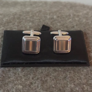 Silvriga manschettknappar från Dako - Stilrena fyrkantiga manschettknappar från Dako i blankt silverfärgat material. De har en klassisk design med diskreta ränder och passar perfekt till skjortor när du vill addera en elegant touch till din outfit.