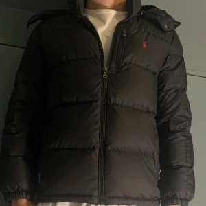 Svart pufferjacka från Polo Ralph Lauren - Svart pufferjacka från Polo Ralph Lauren med huva och dragkedja framtill. Jackan har två sidofickor med dragkedja och den klassiska röda Polo-loggan på bröstet. Perfekt för kalla dagar och har en schysst, lite boxig passform.