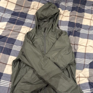 Grön vindjacka från McKinley med huva - Snygg grön vindjacka från McKinley i storlek medium. Jackan har en praktisk huva med dragsko, meshfoder på insidan och hel dragkedja framtill. Perfekt för blåsiga dagar och outdoor-aktiviteter. Enkel design med elastisk nederkant och diskret logga på huvan.