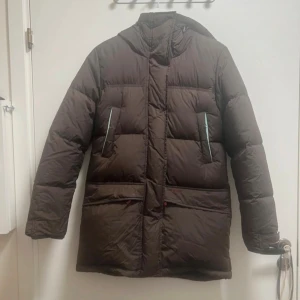 Brun lång dunjacka från peak performance  - Säljer en brun, lång dunjacka från peak performance med huva och svarta detaljer på insidan. Jackan har quiltad design, dragkedja och knappar framtill samt praktiska fickor med dragkedja. Perfekt för kalla dagar och har en clean, stilren look.