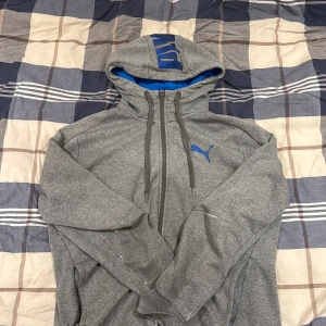 Grå hoodie från Puma med blå detaljer - Säljer en grå hoodie från Puma med blå logga på bröstet och stort blått PUMA-tryck på huvan. Tröjan har dragkedja, snörning i huvan och blått innerfoder. Perfekt för chill eller träning, klassisk och sportig look.