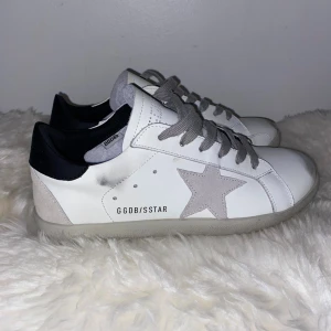 Golden Goose Superstar sneakers vit/grå - Golden Goose Superstar sneakers i vitt skinn med grå mockadetaljer och grå snörning. Ikonisk stjärna på sidan och svart häl med präglad logga. Platt sula och rund tå, insida med Super-Star branding. Snygg och stilren modell som passar till allt.