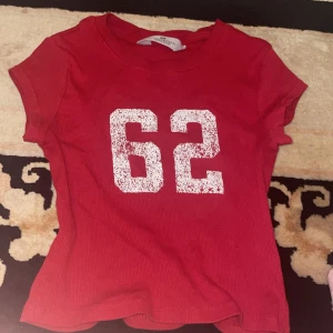 Röd t-shirt med tryck från H&M - Säljer en röd t-shirt från H&M med vitt tryck '62' på framsidan. T-shirten har korta ärmar och jag har haft på mig den 2-3 gånger.