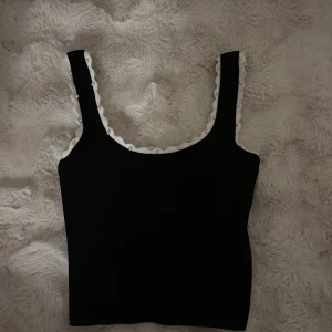 Svart linnetopp med pärldetaljer - Snygg svart linnetopp med vit vågig kant och små pärlor längs urringningen och axelbanden. Toppen är ärmlös och har en klassisk, rak form som passar perfekt till jeans eller kjol. Perfekt för dig som gillar stilrena detaljer och vill sticka ut lite extra.