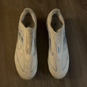 Adidas F50 vita fotbollsskor - Säljer ett par Adidas F50 fotbollsskor i vit färg med blå detaljer och mönster. Skorna har en strömlinjeformad design, platt sula med dobbar för gräsplan och är tillverkade i syntetmaterial. Perfekta för dig som vill ha lättviktsskor med grym känsla på planen.  Box följer ej med 